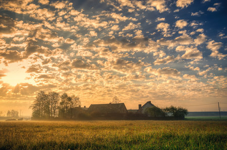 Spring rural landscape. Old houses and morning fog in the meadow with clouds and sun in the background.の写真素材