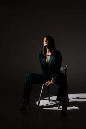 Studio photo of pretty brunette woman in twilight sitting on black chair. A ray of light hits her face.の写真素材