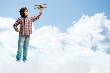 Boy in helmet pilot playing with a toy wooden airplane in the clouds, dreaming of becoming a pilotの写真素材