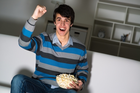 young man watching television with popcorn, raised his hand and cheering for their teamの写真素材