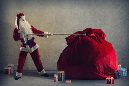 Santa Claus pulls a huge bag of gifts lying around boxes with giftsの写真素材