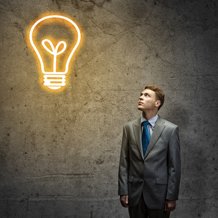 Young thoughtful businessman looking above at light bulbの写真素材