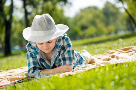 Cute boy in summer park lying on blanket and reading bookの写真素材
