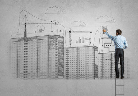 Rear view of businessman standing on ladder and drawing buildingsの写真素材