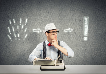 Young guy writer in hat and glasses using typing machineの写真素材
