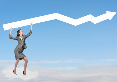 Young businesswoman standing on cloud holding white arrowの写真素材