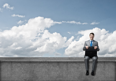 Young businessman with suitcase sitting on roof of buildingの写真素材