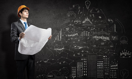 Young thoughtful man engineer in helmet with project in hands and sketches on wallの写真素材