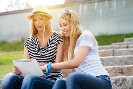 Two young female friends sitting on steps and using tablet pcの写真素材