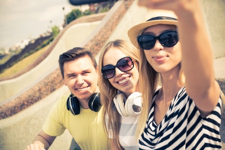 Three young happy people sitting outdoors and making selfieの写真素材