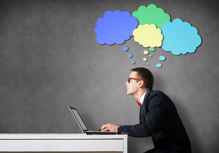 Young funny man wearing glasses sitting at table and working on laptopの写真素材