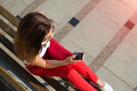 Top view of young pretty brunette with mobile phone sitting on bench in summer parkの写真素材