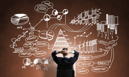 Rear view of businessman sitting in office chair and looking at infographs on chalkboardの写真素材