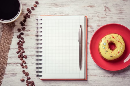 Opened notepad and cup of coffee with donut on wooden tableの写真素材