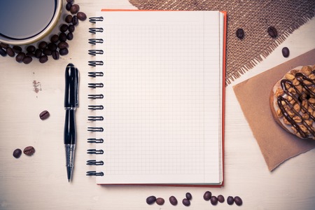 Opened notepad and cup of coffee with donut on wooden tableの写真素材