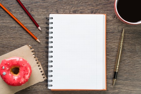 Opened notepad and cup of coffee with donut on wooden tableの写真素材