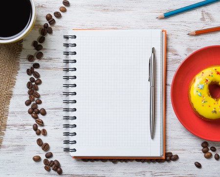 Opened notepad and cup of coffee with donut on wooden tableの写真素材