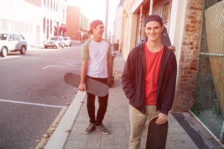 Positive teenage friends with skateboards walking down the street in summer dayの写真素材