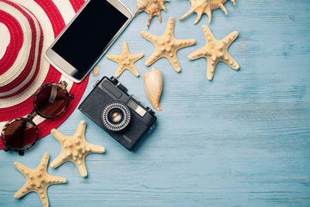Straw hat sunglasses smartphone and photocamera among sea shells and stones on wooden surfaceの写真素材