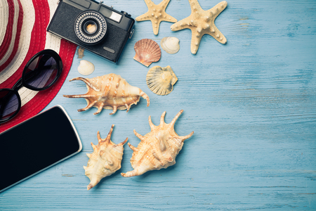 Straw hat sunglasses smartphone and photocamera among sea shells and stones on wooden surfaceの写真素材