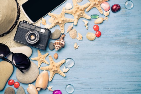 Straw hat sunglasses smartphone and photocamera among sea shells and stones on wooden surfaceの写真素材