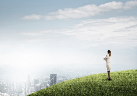 Businesswoman standing with back on top of hill and viewing modern cityの写真素材
