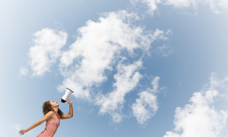 Young woman shouting in megaphone with blue sky backgroundの写真素材