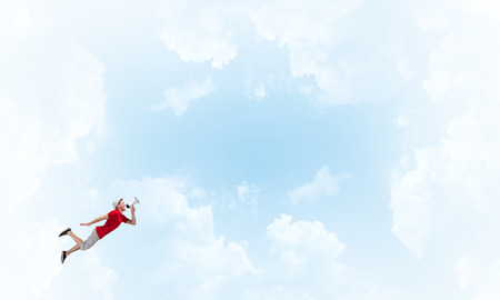 Young cheerful man with megaphone flying high in skyの写真素材