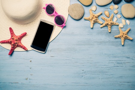 Straw hat sunglasses smartphone among sea shells and stones on wooden surfaceの写真素材