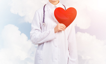 Young woman doctor against sky background holding red heartの写真素材