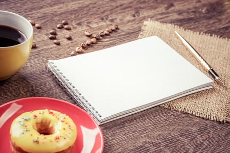 Opened notepad and cup of coffee with donut on wooden tableの写真素材