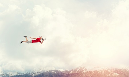 Young cheerful man with megaphone flying high in skyの写真素材