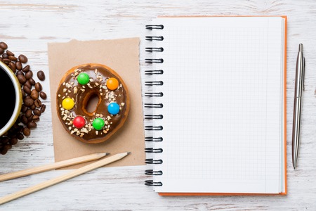 Opened notepad and cup of coffee with donut on wooden tableの写真素材