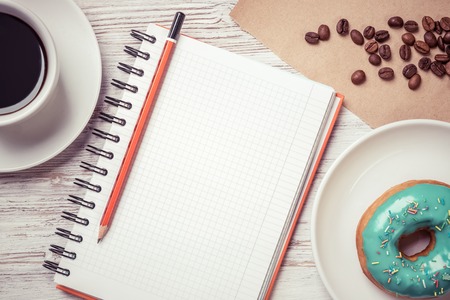 Opened notepad and cup of coffee with donut on wooden tableの写真素材