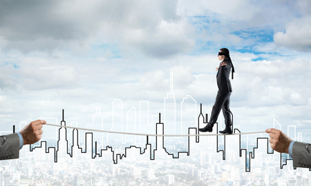 Businessman with blindfolder on eyes walking on rope over cityscape backgroundの写真素材