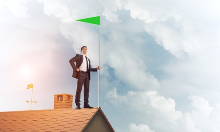 Businessman standing on house roof and holding green flag. Mixed mediaの写真素材
