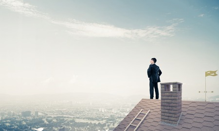 Young determined businessman standing on house roof and looking away. Mixed mediaの写真素材