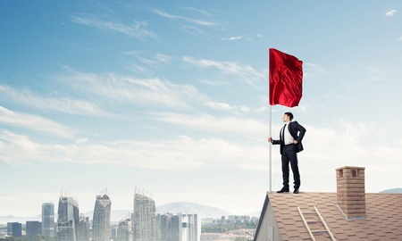 Businessman standing on house roof and red holding flag. Mixed mediaの写真素材