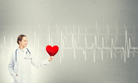 Young woman doctor against gray background holding red heartの写真素材