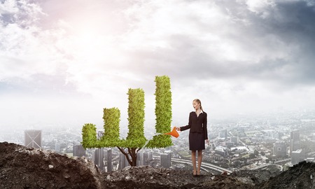 Young attractive businesswoman watering symbol chart plant in ground with can against the backdrop of the cityの写真素材