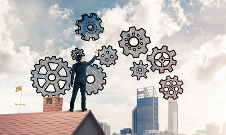 Young businessman standing on house roof and drawing gear mechanism. Mixed mediaの写真素材
