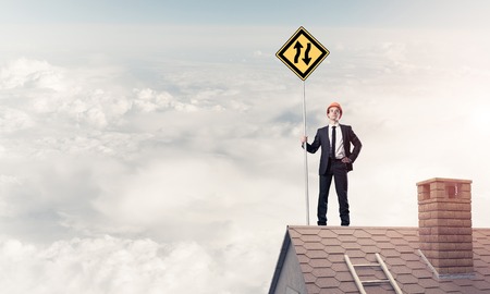 Young determined businessman standing on house roof and showing yellow roadsign. Mixed mediaの写真素材