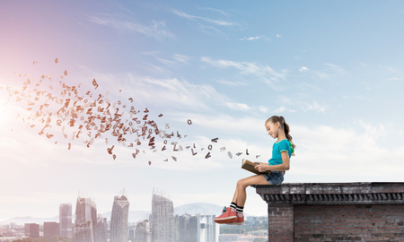 Cute kid girl sitting on building roof reading book and letters fly in airの写真素材