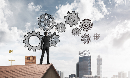 Young businessman standing on house roof and drawing gear mechanism. Mixed mediaの写真素材