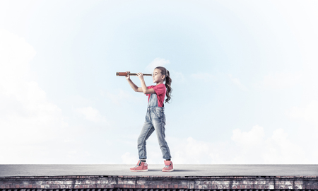 Cute kid girl standing on house roof and looking in spyglassの写真素材