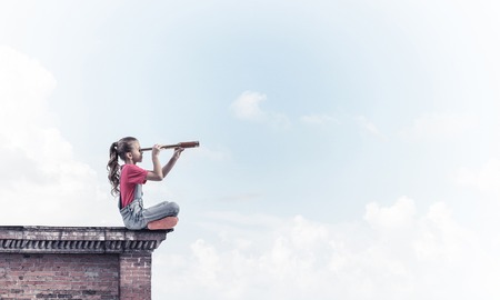 Cute kid girl sitting on house roof and looking in spyglassの写真素材
