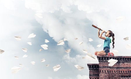 Cute kid girl sitting on house roof and looking in spyglassの写真素材