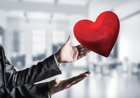 Cropped image of business woman in suit keeping big red heart in his hands with office view and sunlight on background. Mixed media.の写真素材