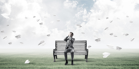 Young shocked businessman sitting on wooden bench and covering eyes with palmsの写真素材