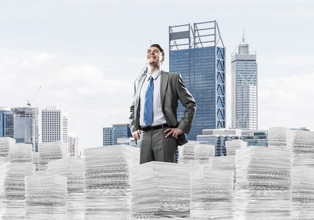 Confident businessman in suit standing on pile of documents and looking away with cityscape on background. Mixed media.の写真素材
